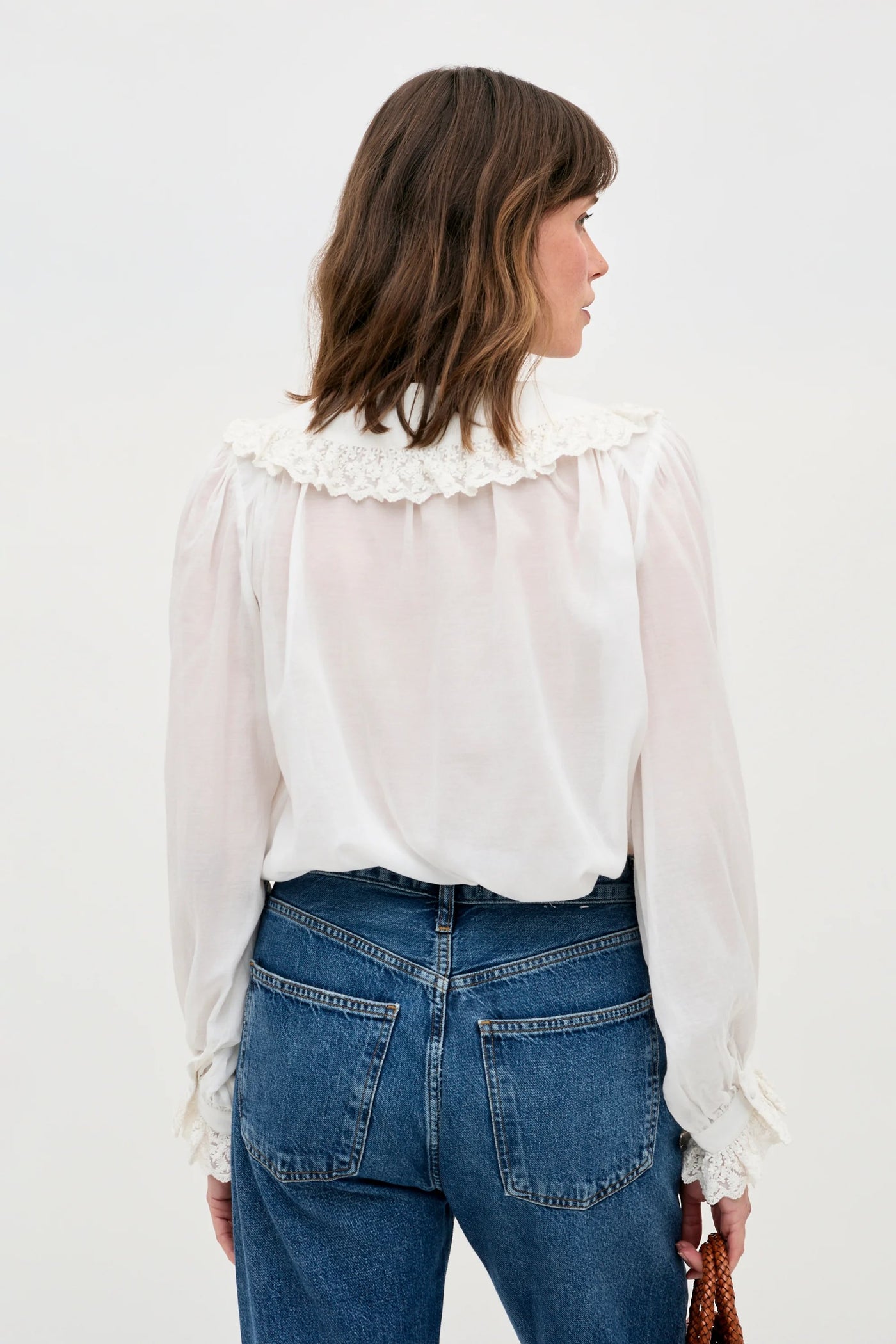 A woman with shoulder-length brown hair, seen from behind, wears the Milia - White blouse, featuring lace details on the collar and cuffs, paired with high-waisted blue jeans. She carries a brown woven handbag.