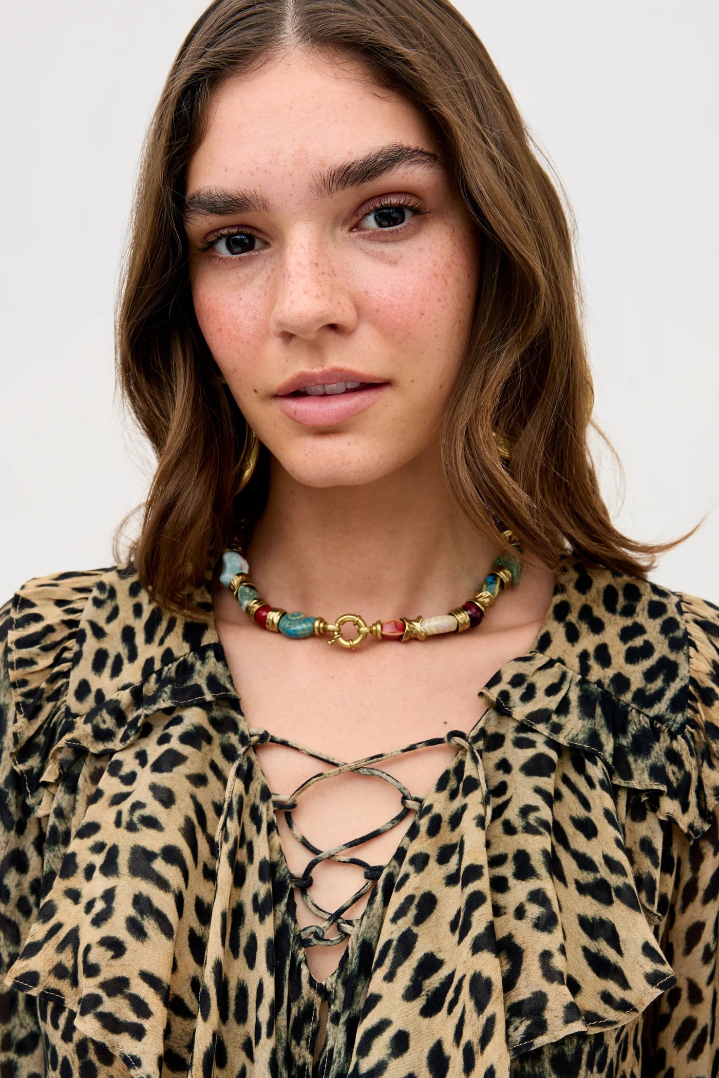 A woman with wavy brown hair wears the Milia - Nina Leopard Brown lace-up top and a chunky, colorful bead necklace. She faces forward, looking slightly serious, against a plain light background.