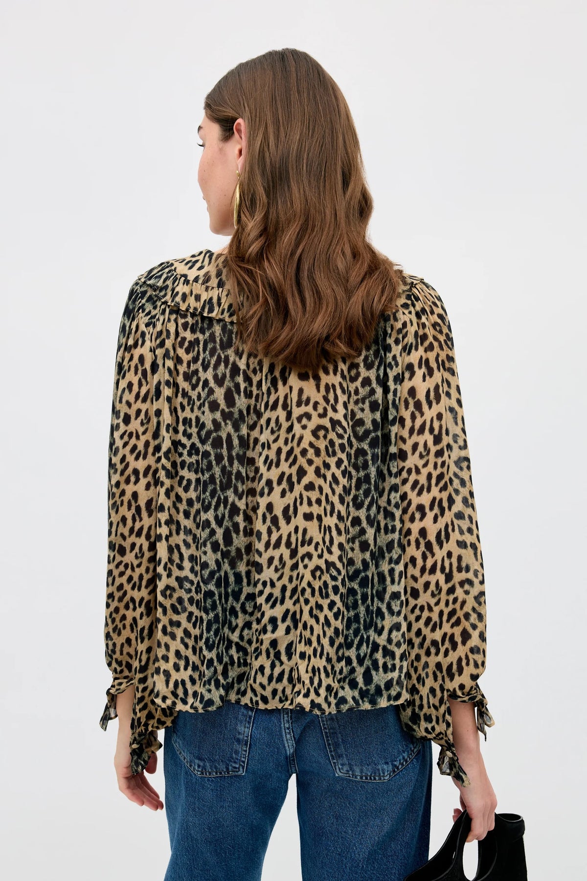A woman with wavy brown hair, seen from behind, wears the Milia - Nina Leopard Brown blouse—a loose, long-sleeve leopard print top—paired with blue jeans and holding a black bag. The background is plain white.