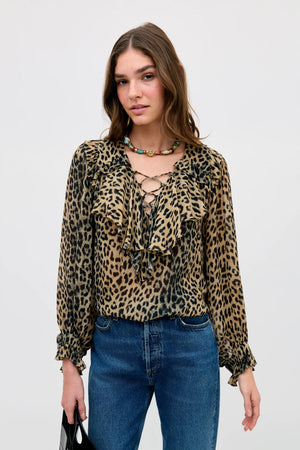A woman stands against a plain light background wearing the Milia - Nina Leopard Brown blouse with ruffles and a lace-up front, paired with blue jeans and a beaded necklace. She holds a black bag.