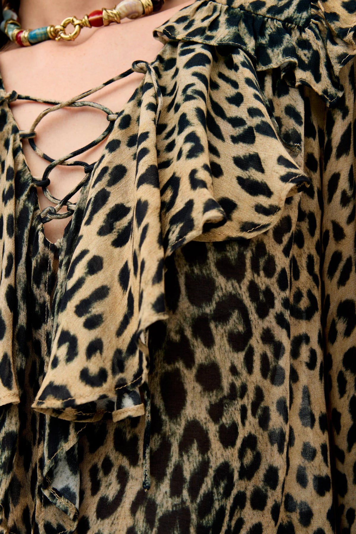 Close-up of a person wearing the Milia - Nina Leopard Brown, a sheer leopard-print blouse featuring ruffled detailing and crisscross lacing at the neckline, paired with a beaded necklace.
