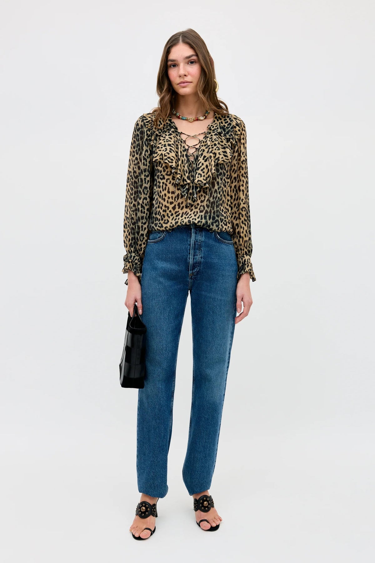 A woman stands against a white background wearing the Milia - Nina Leopard Brown blouse, paired with blue high-waisted jeans, black heeled sandals, a chunky necklace, and holding a small black handbag.