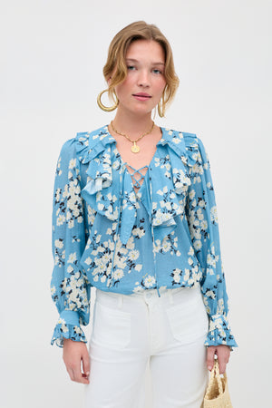 A woman poses against a plain background in the Milia - Amelia Bouquet Blue blouse and white pants, accessorized with large hoop earrings, a gold necklace, and a woven bag.
