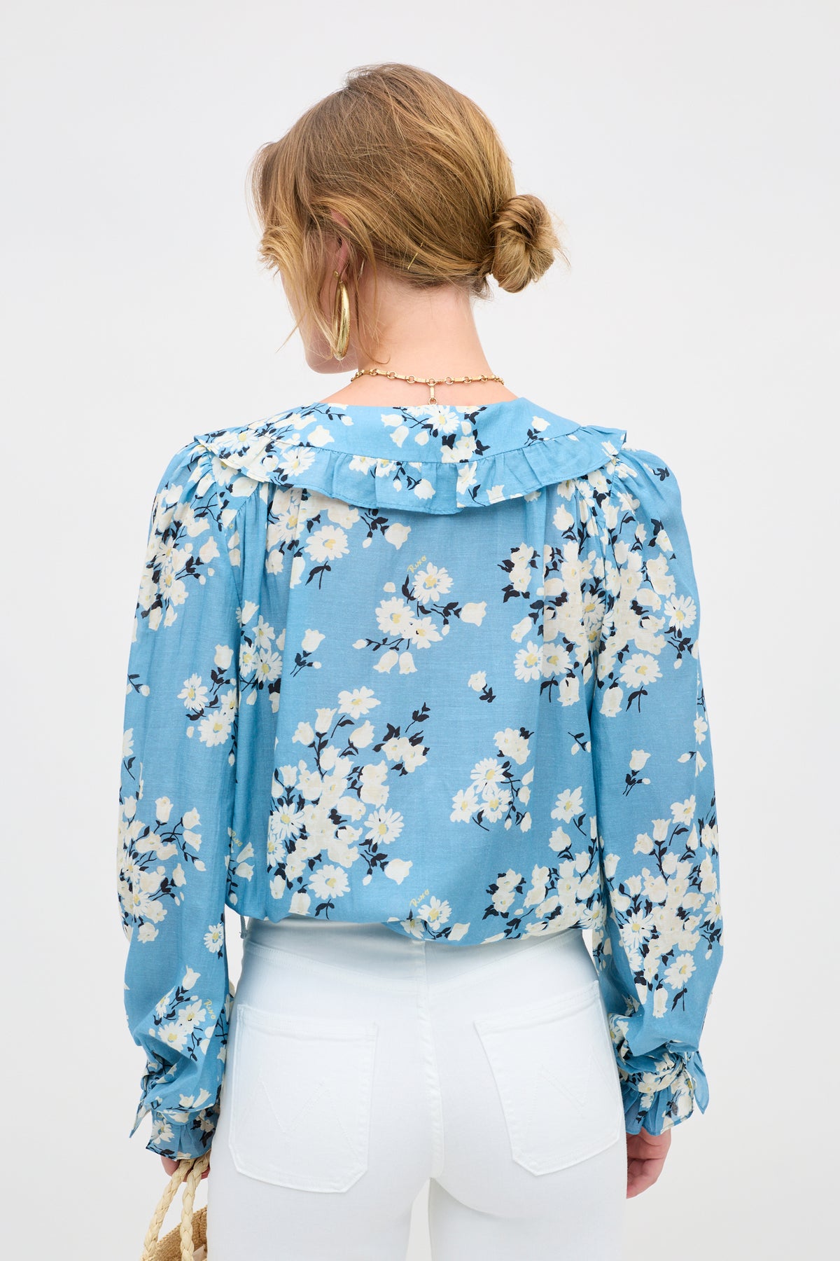 A woman with blonde hair in a low bun, wearing gold hoops and the Milia - Amelia Bouquet Blue blouse, stands with her back to the camera against a plain background, paired with high-waisted white pants.
