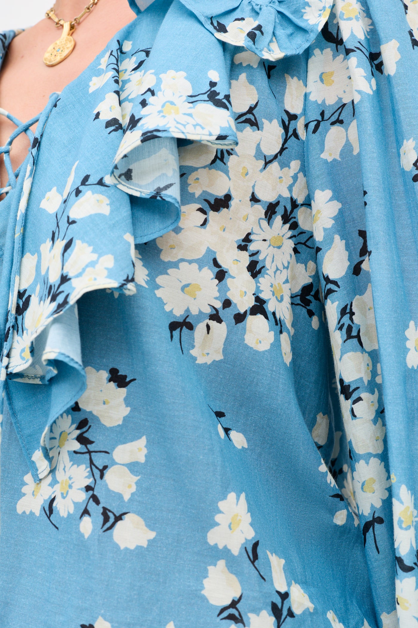 Close-up of the Milia - Amelia Bouquet Blue dress, featuring blue fabric with white floral patterns and ruffled details. A gold necklace with a round pendant appears partially in the top left corner.