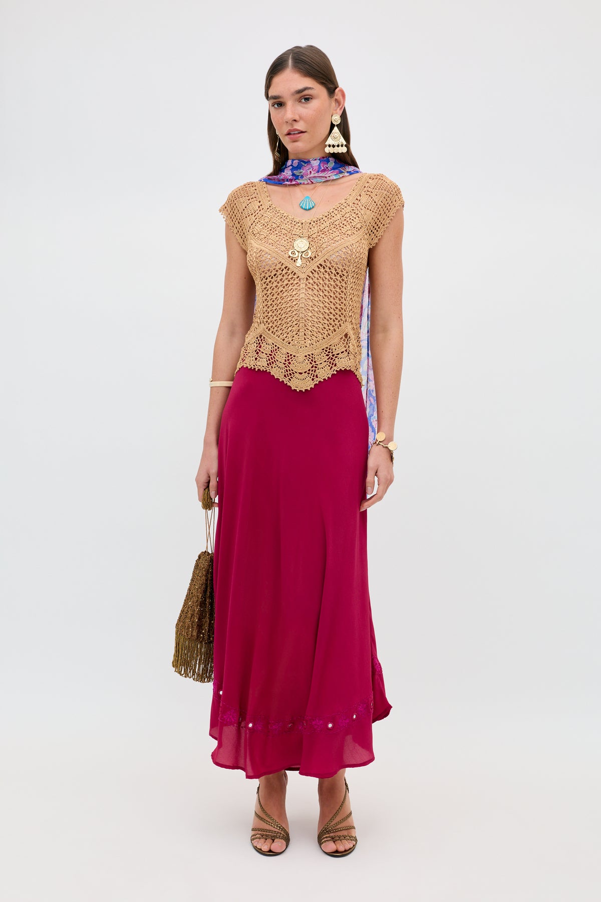 Woman wearing the Kimbra - Gold, a tan crochet top, paired with a long magenta skirt, strappy heels, ornate jewelry, and a purple patterned scarf. She carries a textured brown bag and stands against a plain white background.