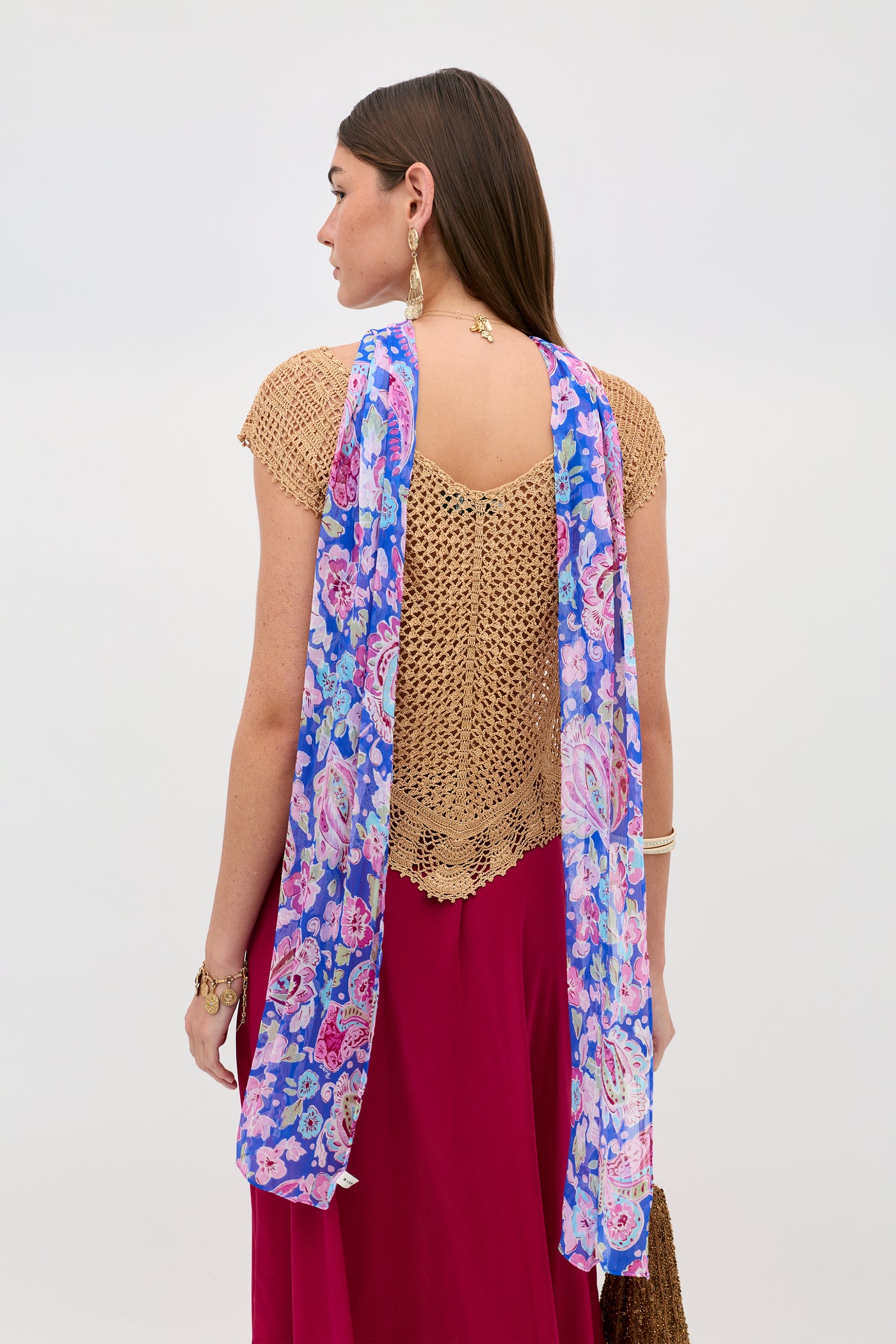 A woman with long brown hair is seen from behind against a plain background, wearing gold Kimbra earrings, a tan crochet top, a pink skirt, and a blue floral scarf draped over her shoulders.