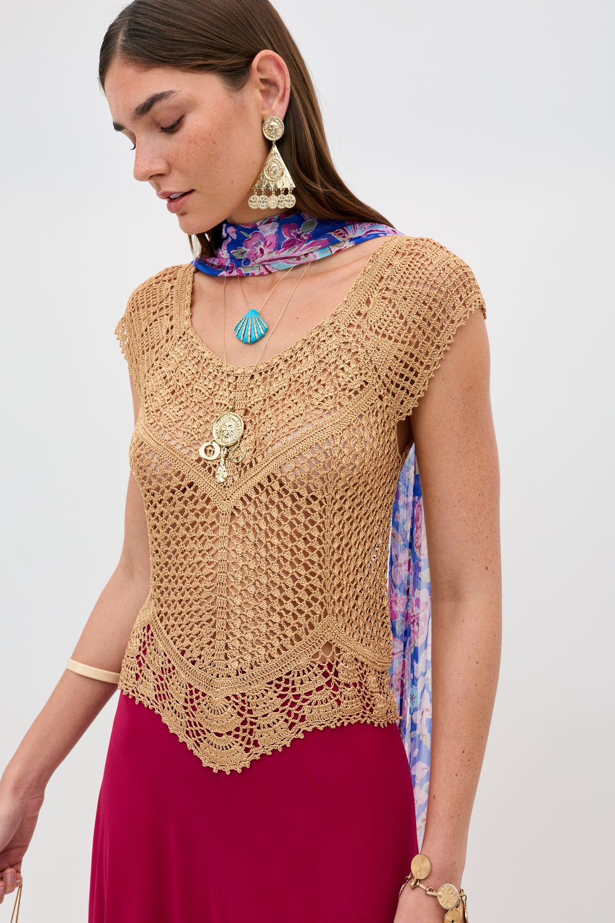 A woman wearing the Kimbra - Gold bracelet stands against a plain background, gazing downward with her arm slightly extended. She is styled in a tan crochet top, red skirt, colorful earrings, layered necklaces, and a purple floral scarf.