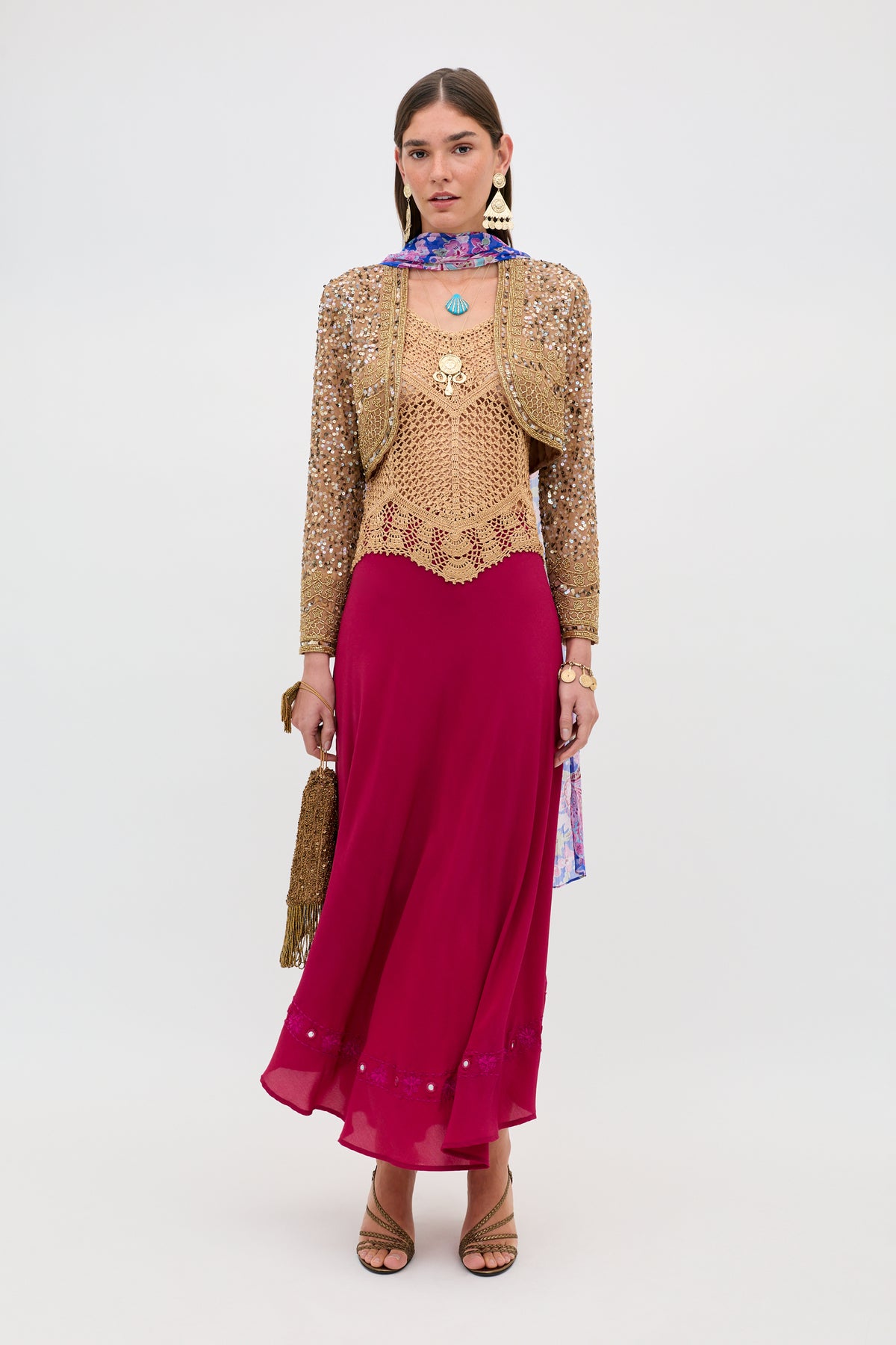 Styled in Kimbra - Gold, she wears a dazzling gold sequin jacket with a beige crochet top, a vibrant magenta skirt, and beige heels. She accessorizes with statement jewelry, a colorful neck scarf, and carries a gold tasseled bag, all posed against a plain background.
