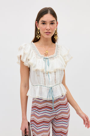 A woman wearing the Melisa - Ivory prairie-inspired ruffle blouse stands against a plain white background, styled with zigzag stripe pants and gold jewelry.