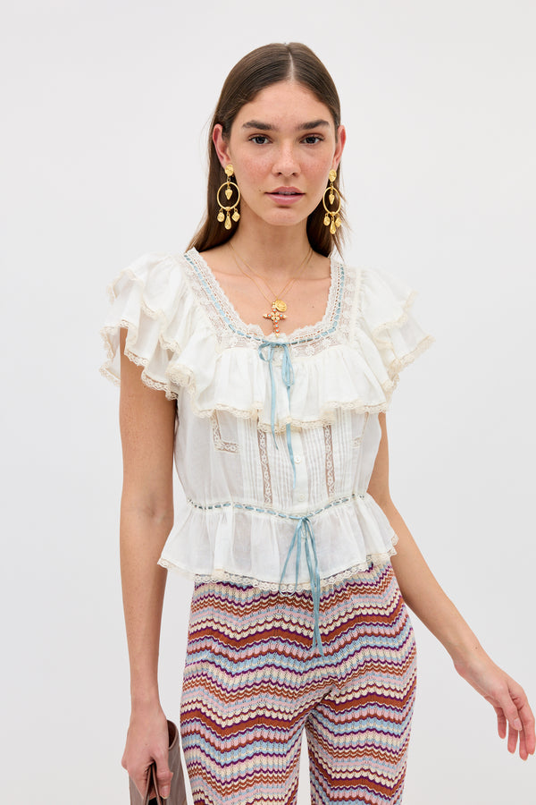 A woman wearing the Melisa - Ivory prairie-inspired ruffle blouse stands against a plain white background, styled with zigzag stripe pants and gold jewelry.