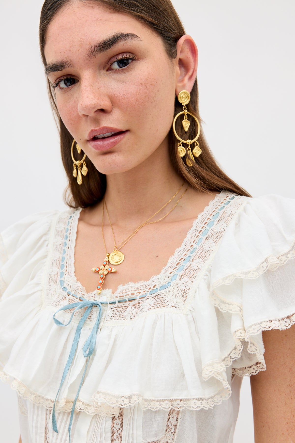 A woman with long brown hair wears Thome - Gold drop earrings, layered gold necklaces, and a white ruffled blouse with lace trim and a blue ribbon bow at the neckline. She gazes neutrally at the camera.