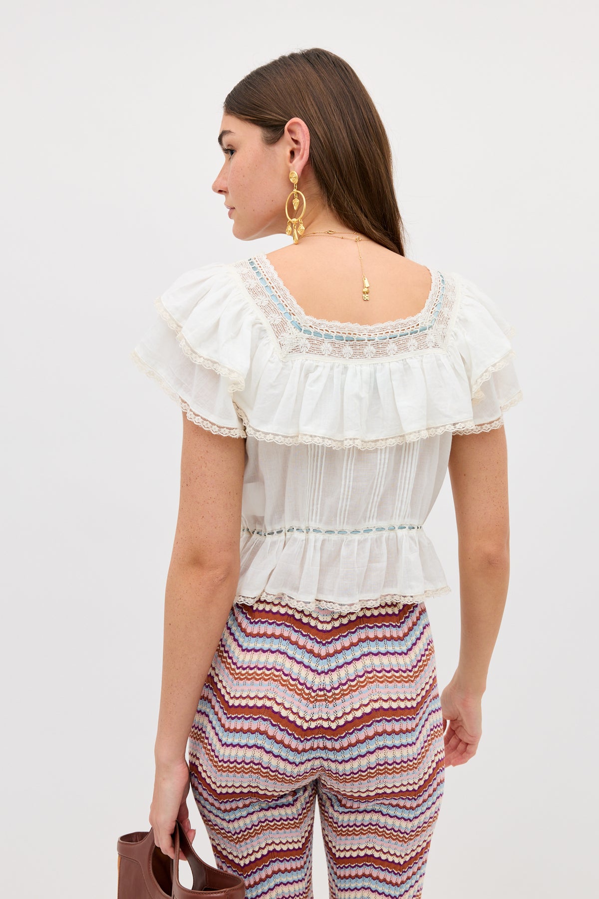 A woman with long brown hair wears the Melisa - Ivory prairie-inspired white cotton ruffle blouse with lace details and colorful zigzag-patterned pants, holding a brown handbag. Turned slightly away, she shows off her earrings and necklace.