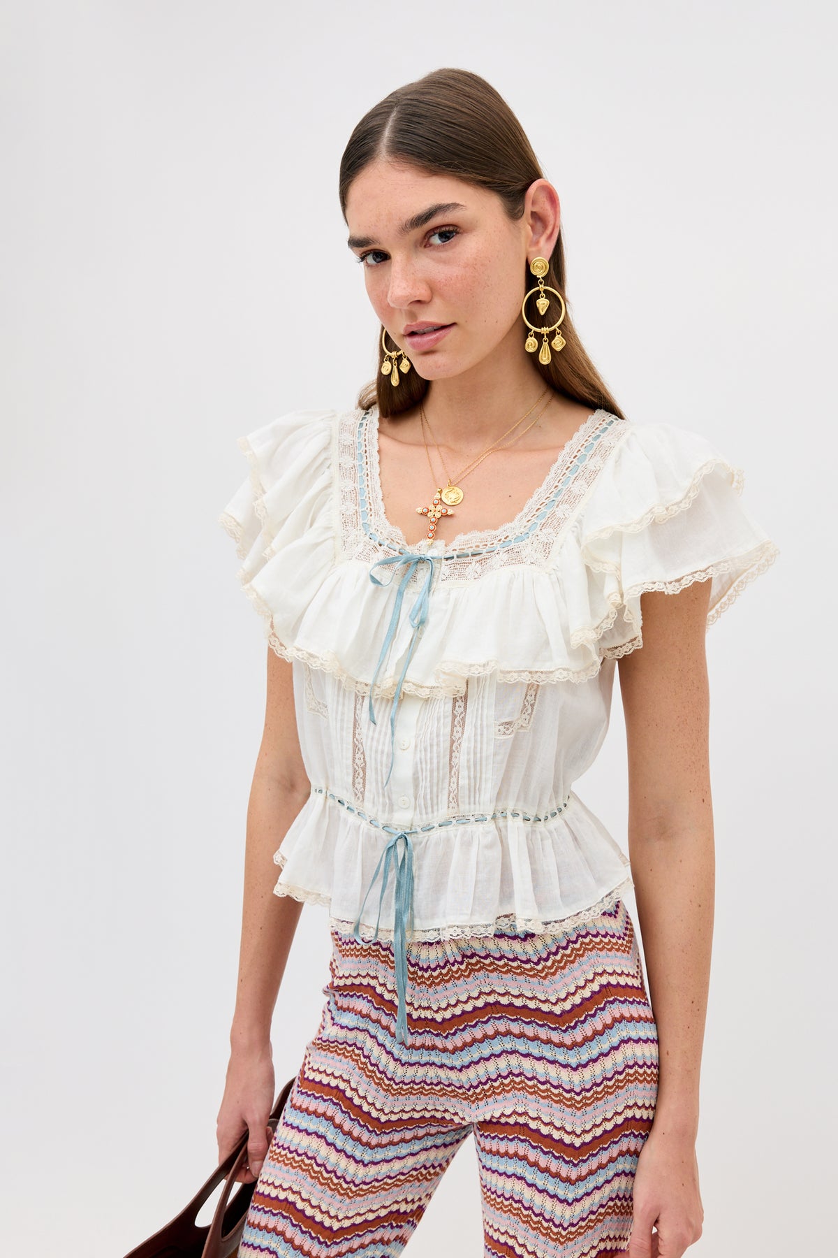 A woman with long brown hair models the Melisa - Ivory prairie-inspired ruffle blouse with lace details and a blue ribbon, paired with patterned multicolor pants, gold earrings, and layered necklaces against a plain light background.