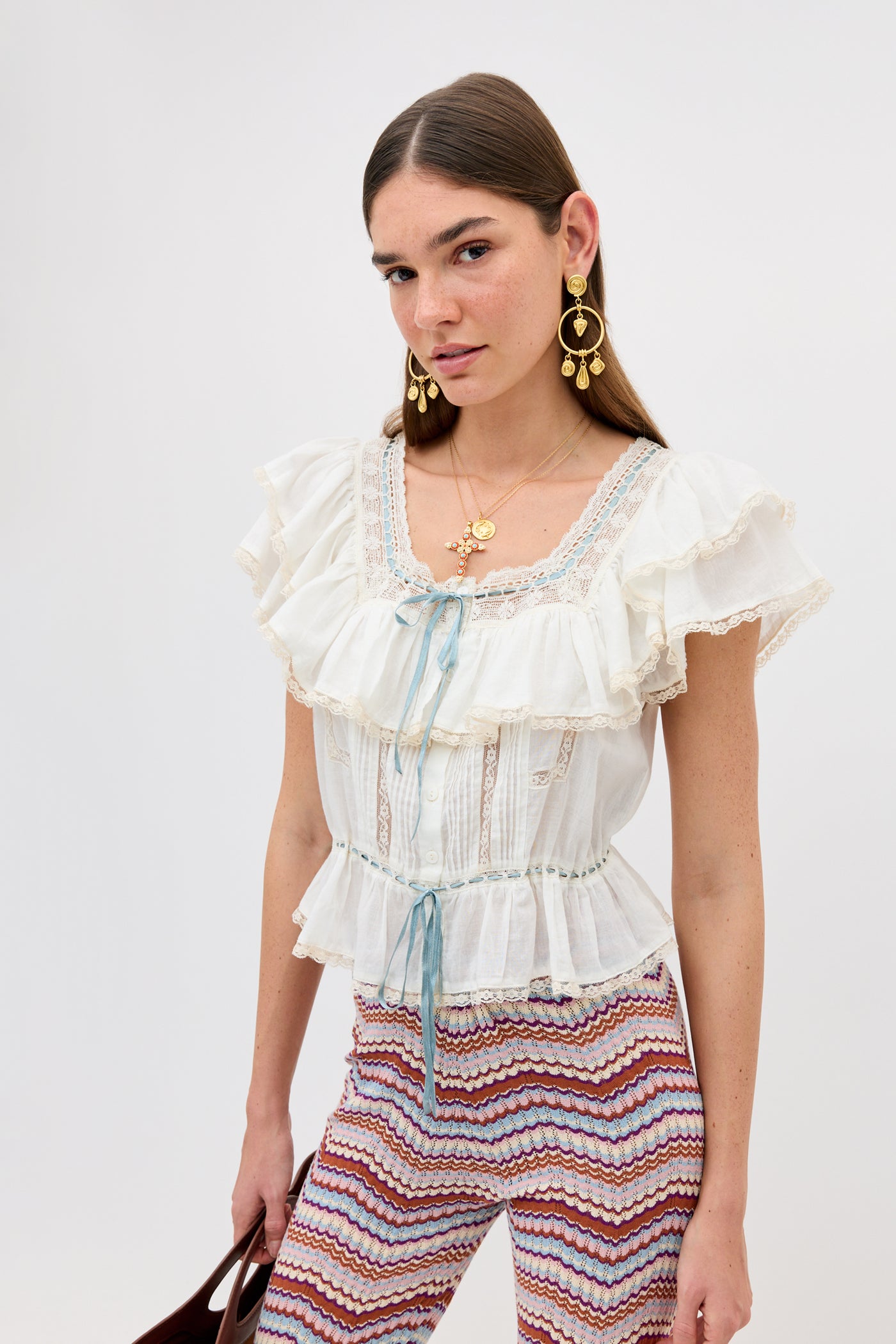 A woman with long brown hair models the Melisa - Ivory prairie-inspired ruffle blouse with lace details and a blue ribbon, paired with patterned multicolor pants, gold earrings, and layered necklaces against a plain light background.