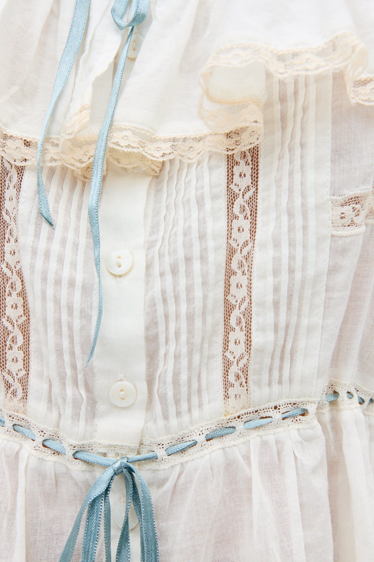 Close-up of the Melisa - Ivory blouse, a prairie-inspired white cotton top with lace trim, vertical lace panels, white buttons, pleats, and light blue ribbon accents. Lightweight semi-sheer fabric adds vintage charm.