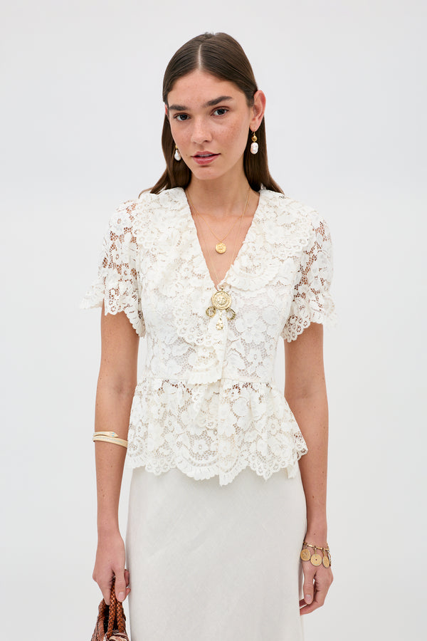 A woman in the Dollie - Cream white lace short-sleeve blouse with a ruffled collar, paired with a cream skirt. She has long brown hair, gold jewelry, and holds a brown bag against a plain white background.