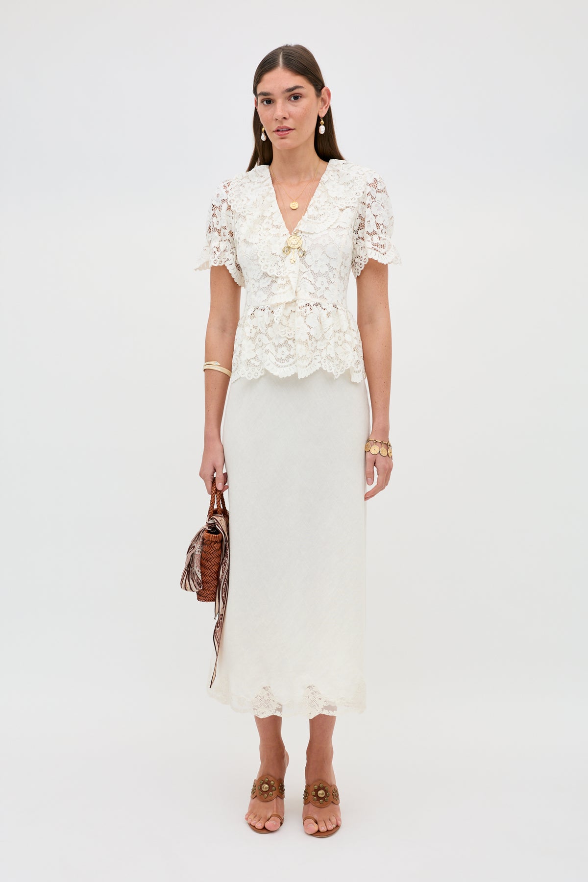 A woman with straight brown hair stands against a plain background, wearing Dollie - Cream: a white lace blouse and matching long skirt. She accessorizes with gold jewelry, brown sandals, and a brown textured handbag.