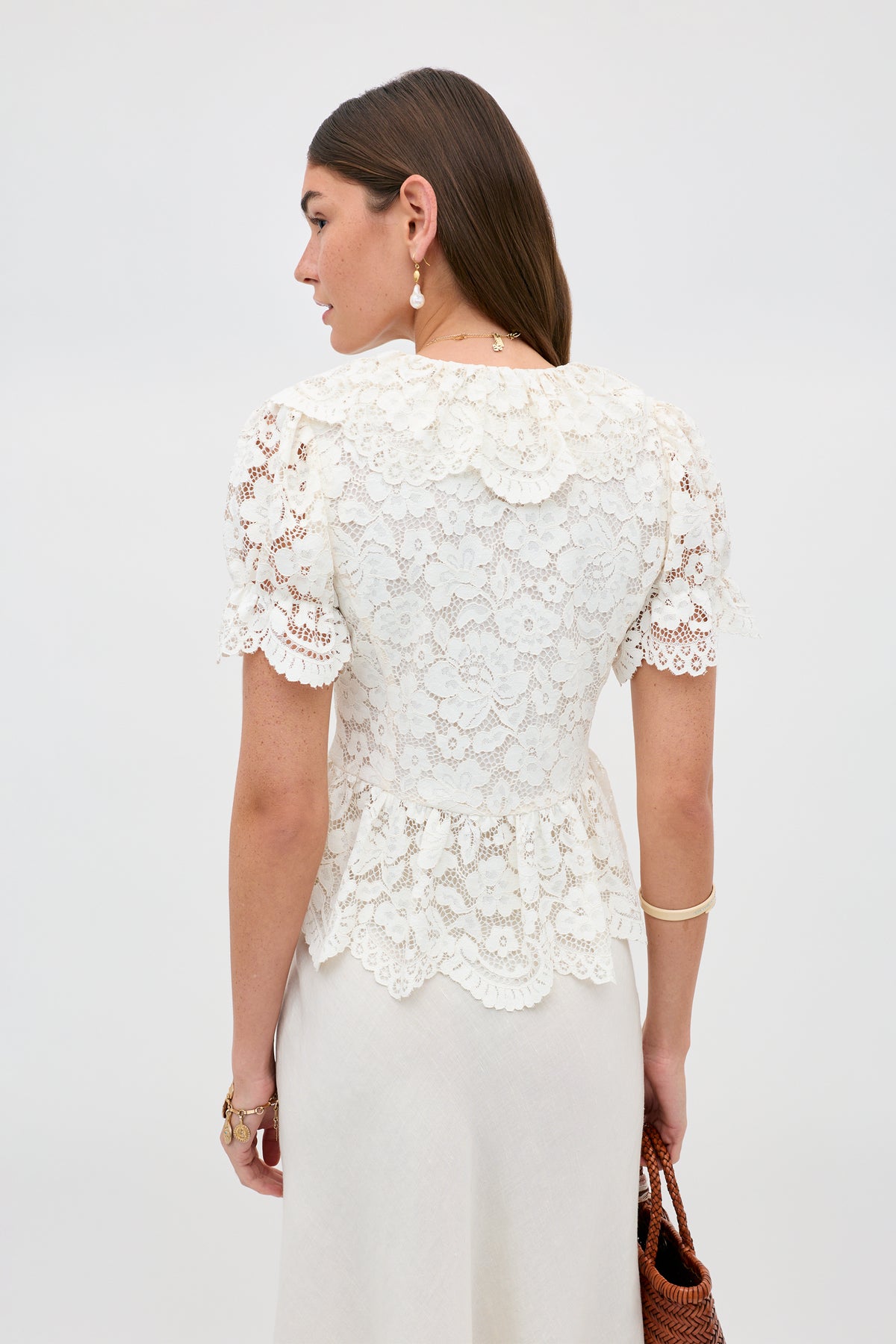 A woman with long brown hair is shown from the back, wearing the Dollie - Cream lace short-sleeve blouse with scalloped edges and a white skirt. She accessorizes with gold jewelry and carries a brown woven bag against a plain white background.