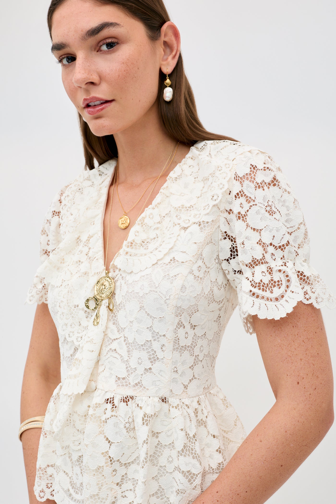 A woman stands against a plain light background wearing Dollie - Cream, a white lace dress with short sleeves, paired with layered gold necklaces, drop earrings, and a bracelet.