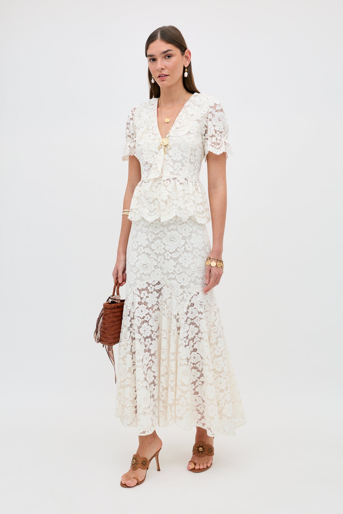A woman stands against a white background wearing the Dollie - Cream lace short-sleeve peplum top with a matching long skirt, tan heeled sandals, gold jewelry, and holding a brown woven handbag.