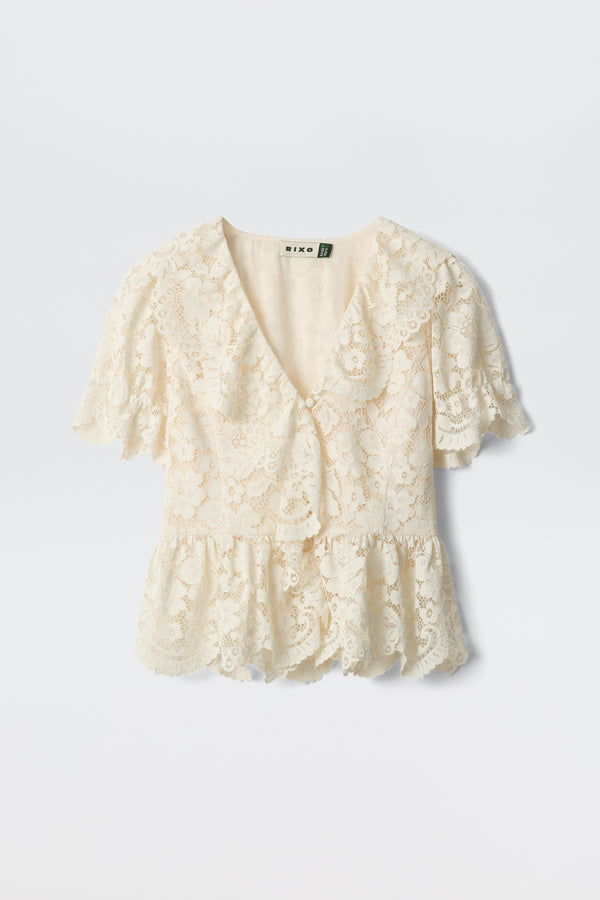 Dollie - Cream: A cream-colored lace blouse featuring short sleeves, a V-neckline, ruffled hem, and scalloped edges, shown against a plain white background.