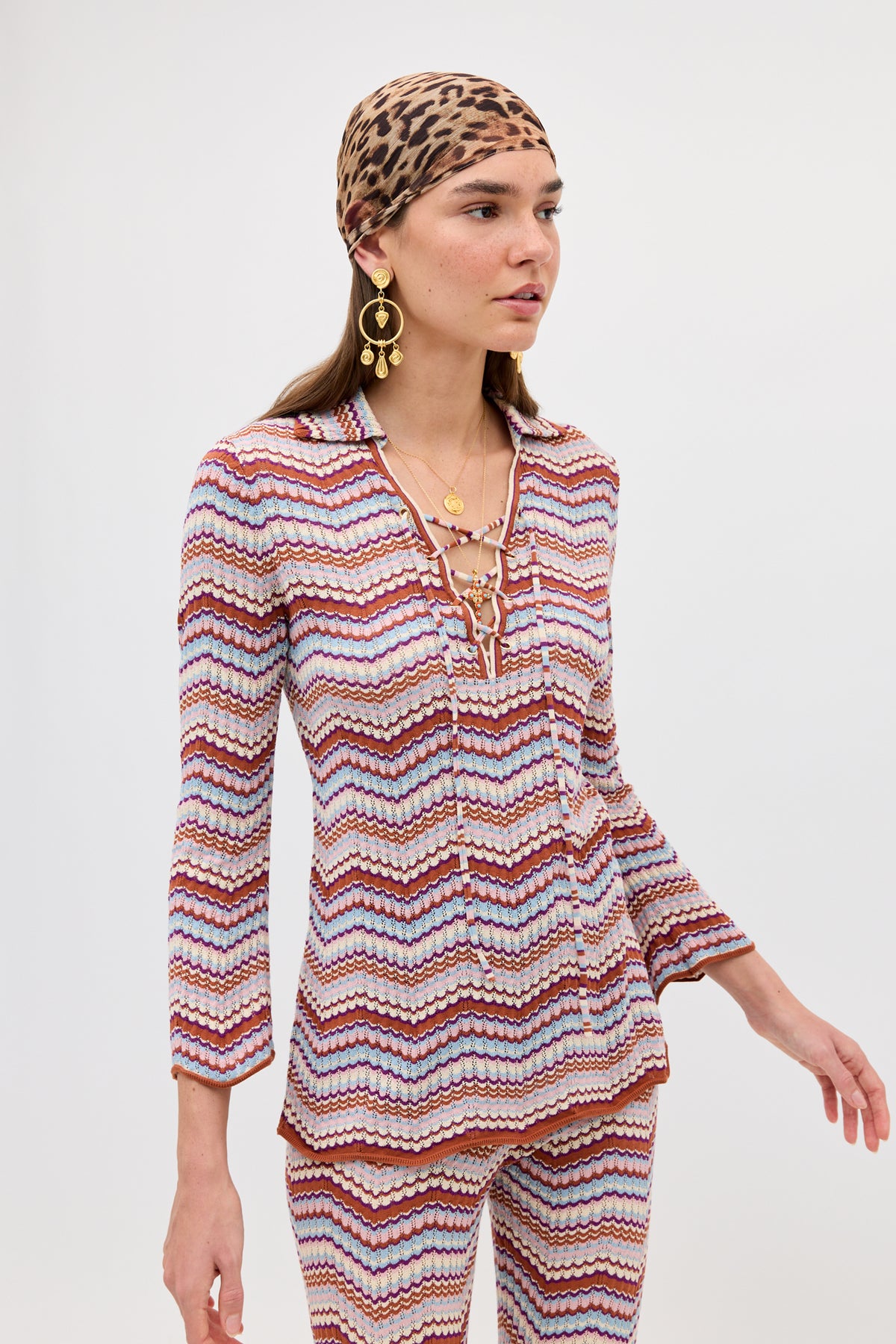 Wearing the Brody - Maria Chevron Multi top with matching pants, a woman stands against a plain white background, styled with large gold earrings, layered necklaces, and a leopard-print headscarf.