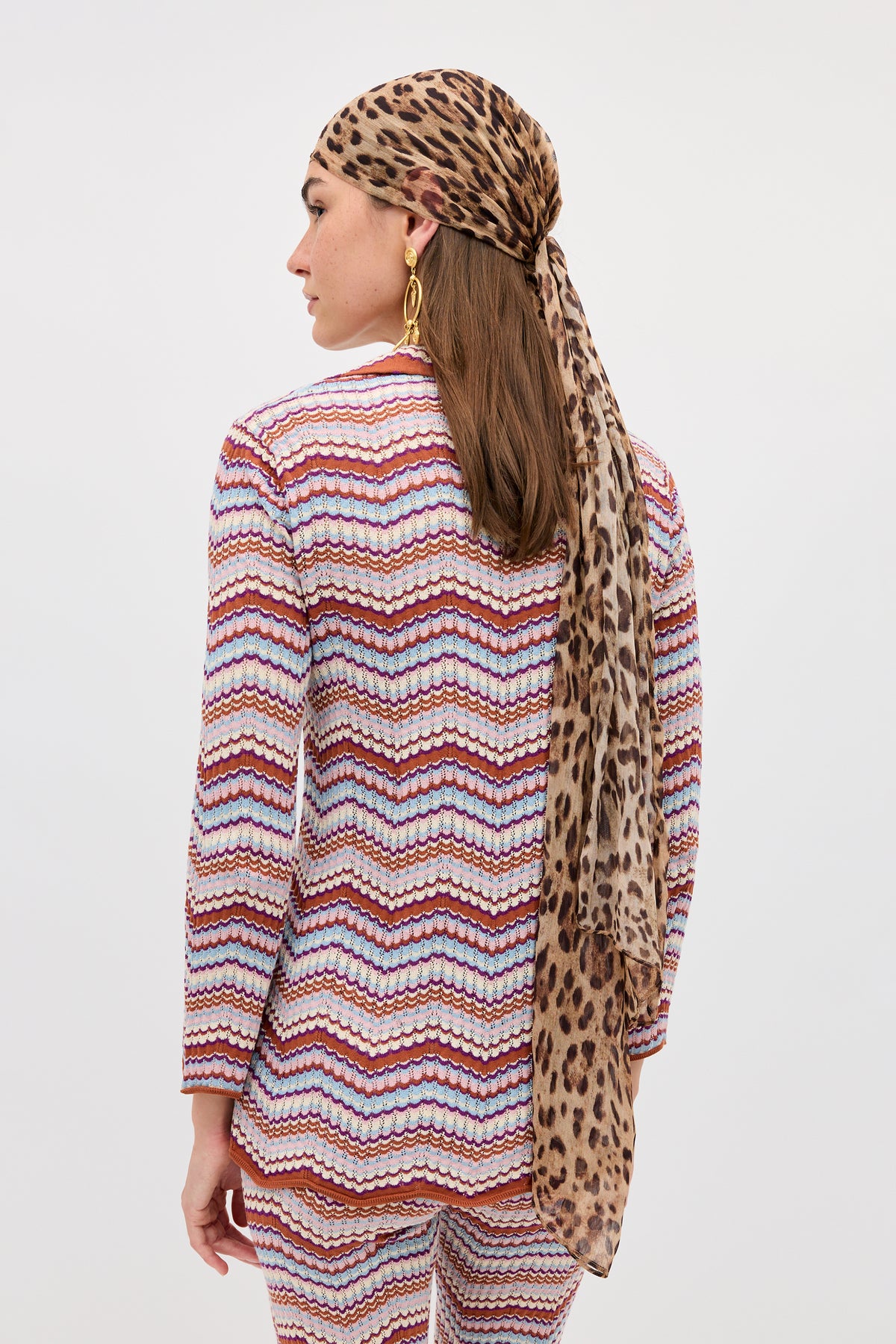 A woman facing away wears the Brody - Maria Chevron Multi suit, which features a vibrant zigzag pattern in pink, white, and red tones. She pairs it with a vintage-inspired top for retro flair, accessorized with a long leopard-print headscarf tied at the back, gold hoop earrings, and her long brown hair cascading down. The background is plain white.