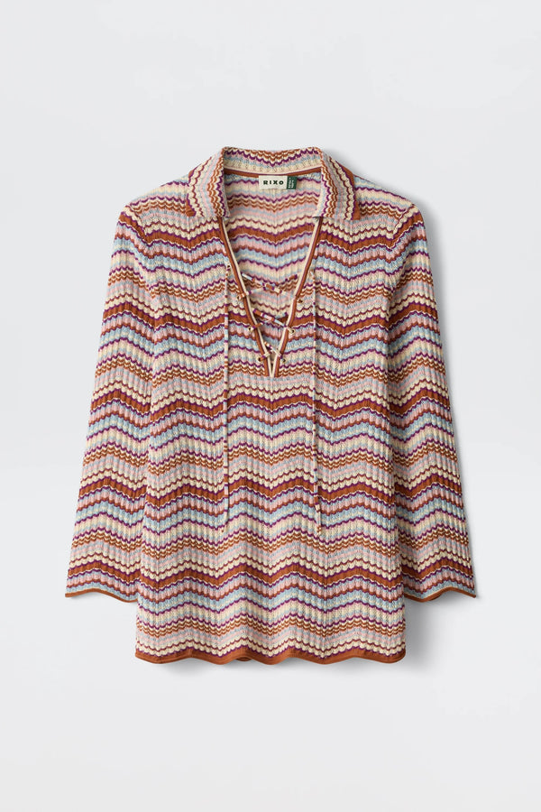 The Brody - Maria Chevron Multi is a vintage-inspired women’s top featuring long sleeves, a collared neckline, and a vibrant pink, purple, and orange chevron pattern. This retro cotton knit piece showcases a charming lace-up V-neck detail and is finished with scalloped edges at the hem and sleeves for an extra touch of playful elegance.