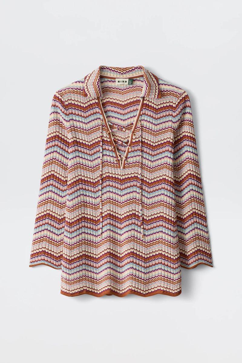 The Brody - Maria Chevron Multi is a vintage-inspired women’s top featuring long sleeves, a collared neckline, and a vibrant pink, purple, and orange chevron pattern. This retro cotton knit piece showcases a charming lace-up V-neck detail and is finished with scalloped edges at the hem and sleeves for an extra touch of playful elegance.