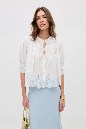A woman wears the erika top ivory, a loose white button-down blouse with lace-trimmed sleeves and hem, paired with a light blue skirt. She accessorizes with a gold bag, gold bracelet, and has wavy brown hair against a plain white background.