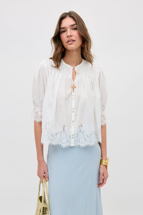 A woman wears the erika top ivory, a loose white button-down blouse with lace-trimmed sleeves and hem, paired with a light blue skirt. She accessorizes with a gold bag, gold bracelet, and has wavy brown hair against a plain white background.