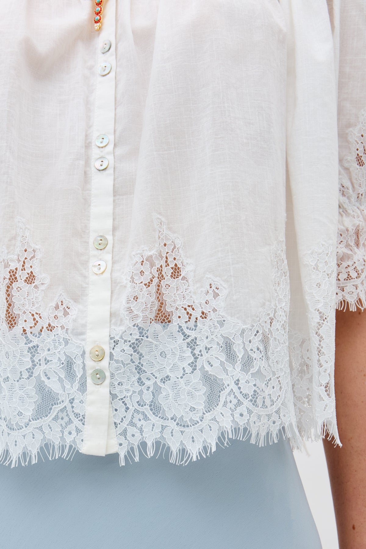 Close-up of the erika top ivory, a white blouse with small buttons and delicate lace trim on the hem and sleeves. The semi-sheer fabric subtly reveals a light blue layer beneath.