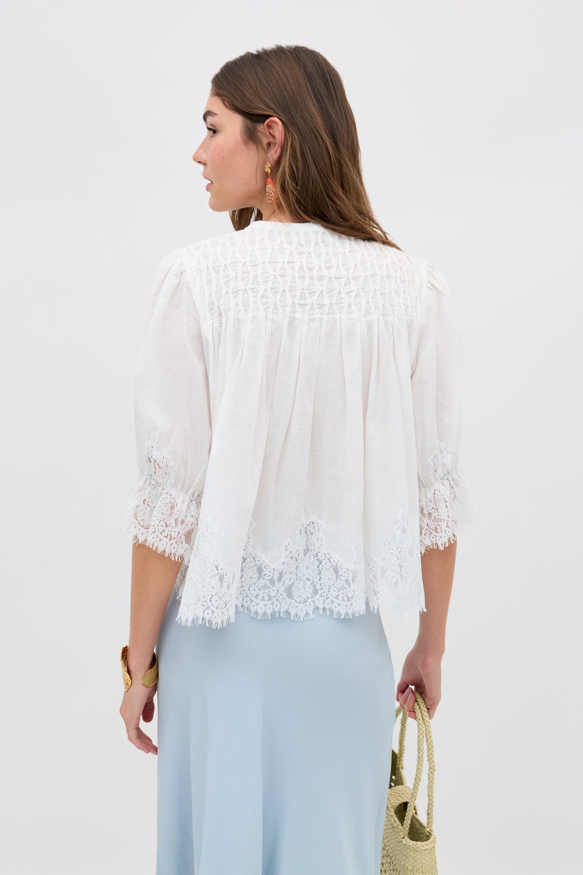 A woman with long brown hair is shown from the side wearing the erika top ivory—a white, lace-trimmed, loose blouse—paired with a light blue skirt and holding a woven straw bag. The detailed back of the top is visible against a plain background.