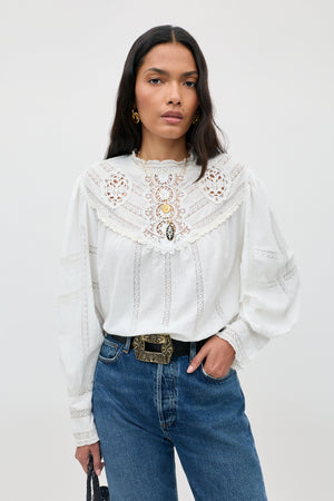 Wearing the Tamzin - Cream blouse, a woman with long dark hair stands against a light gray background, styled with blue jeans, an ornate belt, and gold earrings.