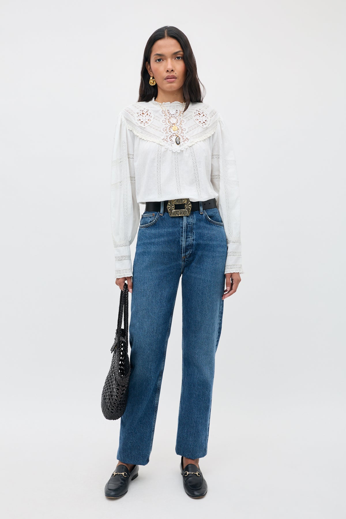 Woman stands against a white background, wearing the Tamzin - Cream long-sleeve blouse with lace details, blue high-waisted jeans, black loafers, a black belt, and holding a black textured handbag.