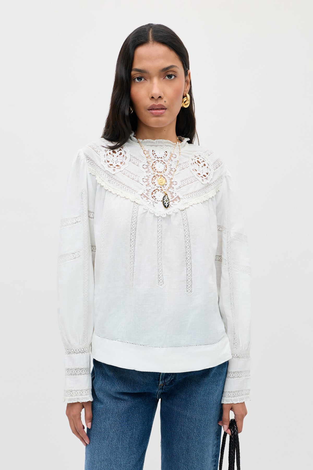 A person with long dark hair wears the Tamzin - Cream blouse with intricate lace detailing, styled with blue jeans and gold earrings, standing against a plain light background while holding a black handbag.