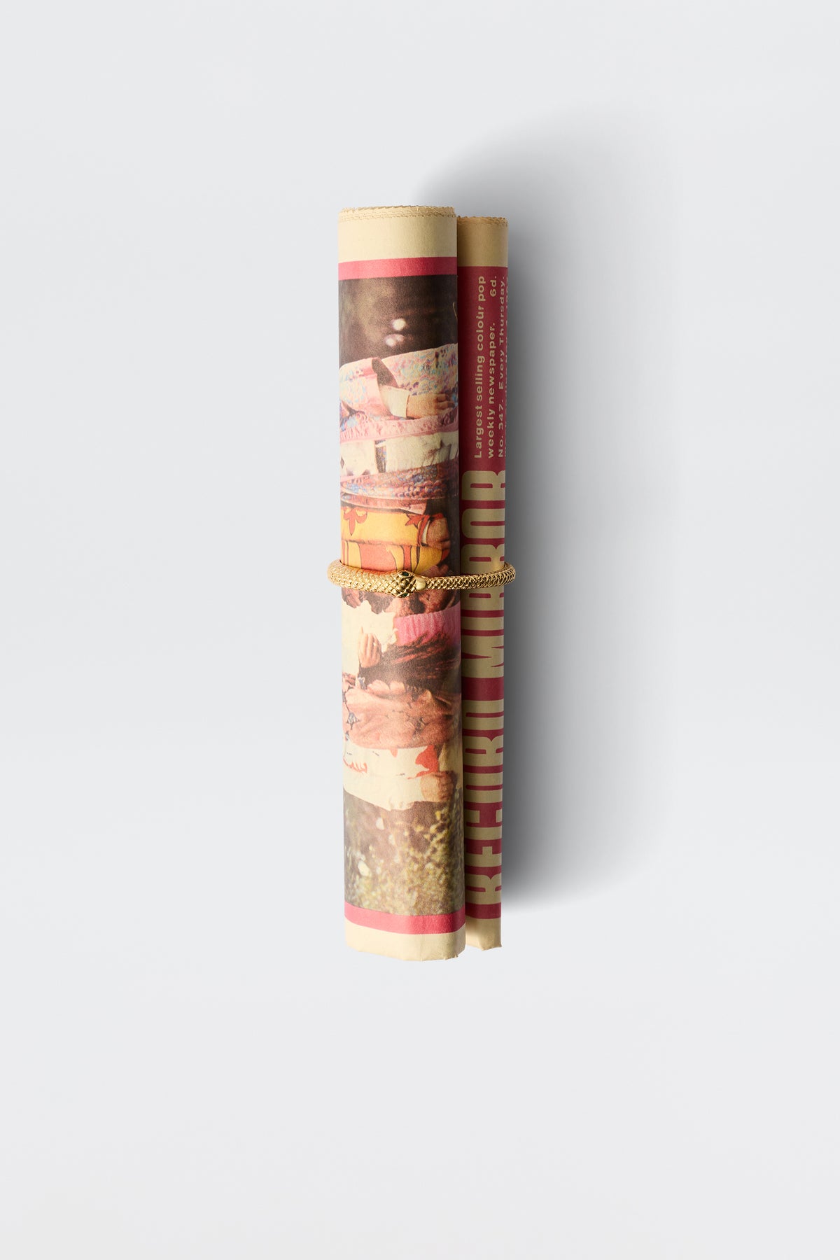The Viera - Gold, a striking gold-plated bracelet with an antique finish, is displayed vertically beside a rolled-up, colorful magazine tied with tan rope against a plain white background.