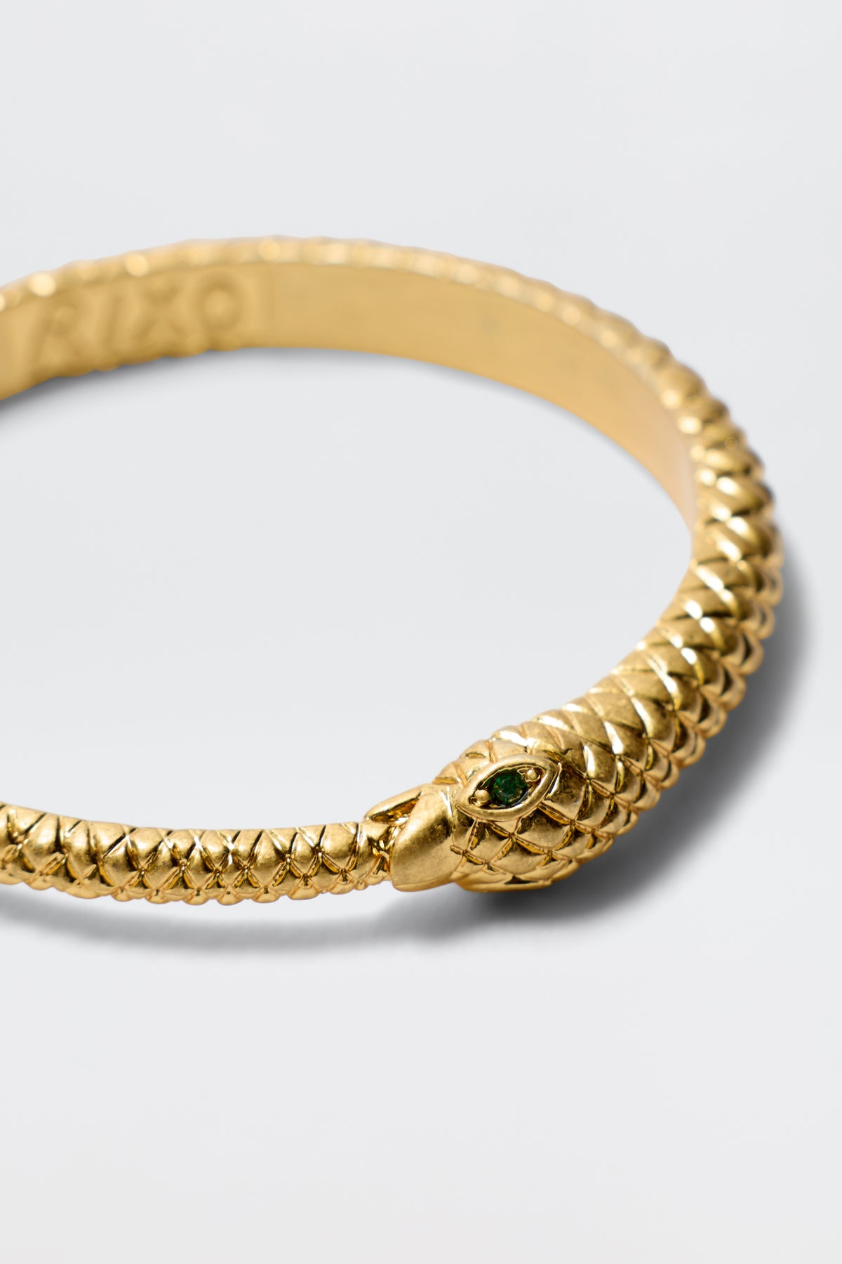 The Viera - Gold is a gold-plated snake bangle with detailed scales, a green gemstone eye, and an antique finish, displayed on a simple light background.