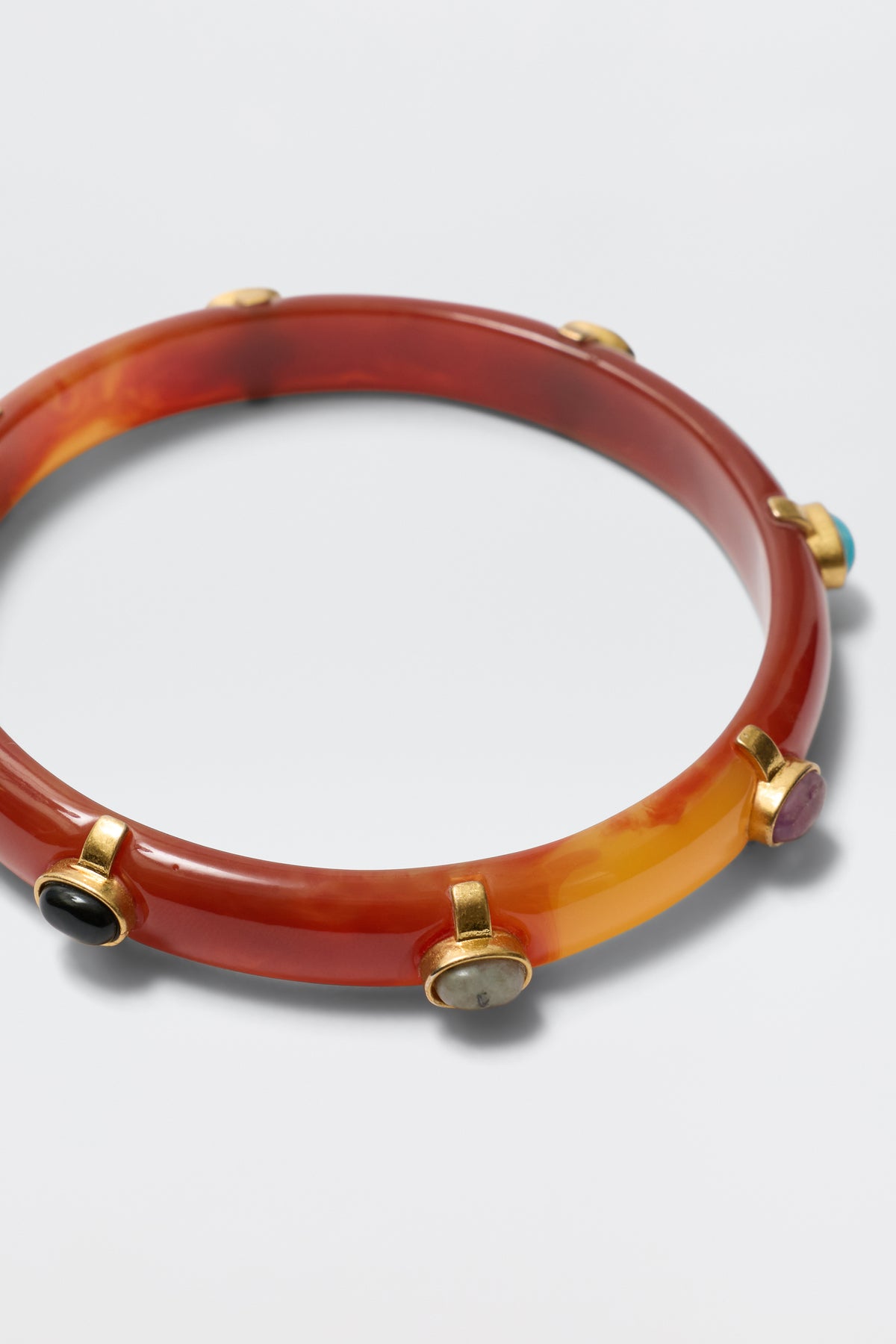 The Sabella - Tortoise is a reddish-brown marbled resin bangle with multicoloured cabochons in gold-plated bezels spaced around its circumference, shown on a white background.