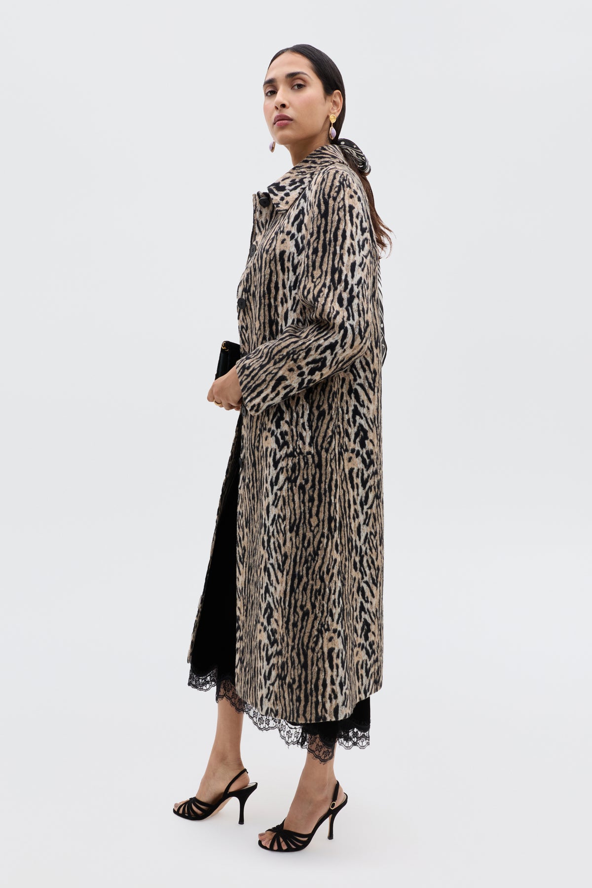 A woman stands against a plain background wearing the Mavi - Black coat over a black lace dress, paired with glamorous sandals, hoop earrings, and holding a clutch by her side.
