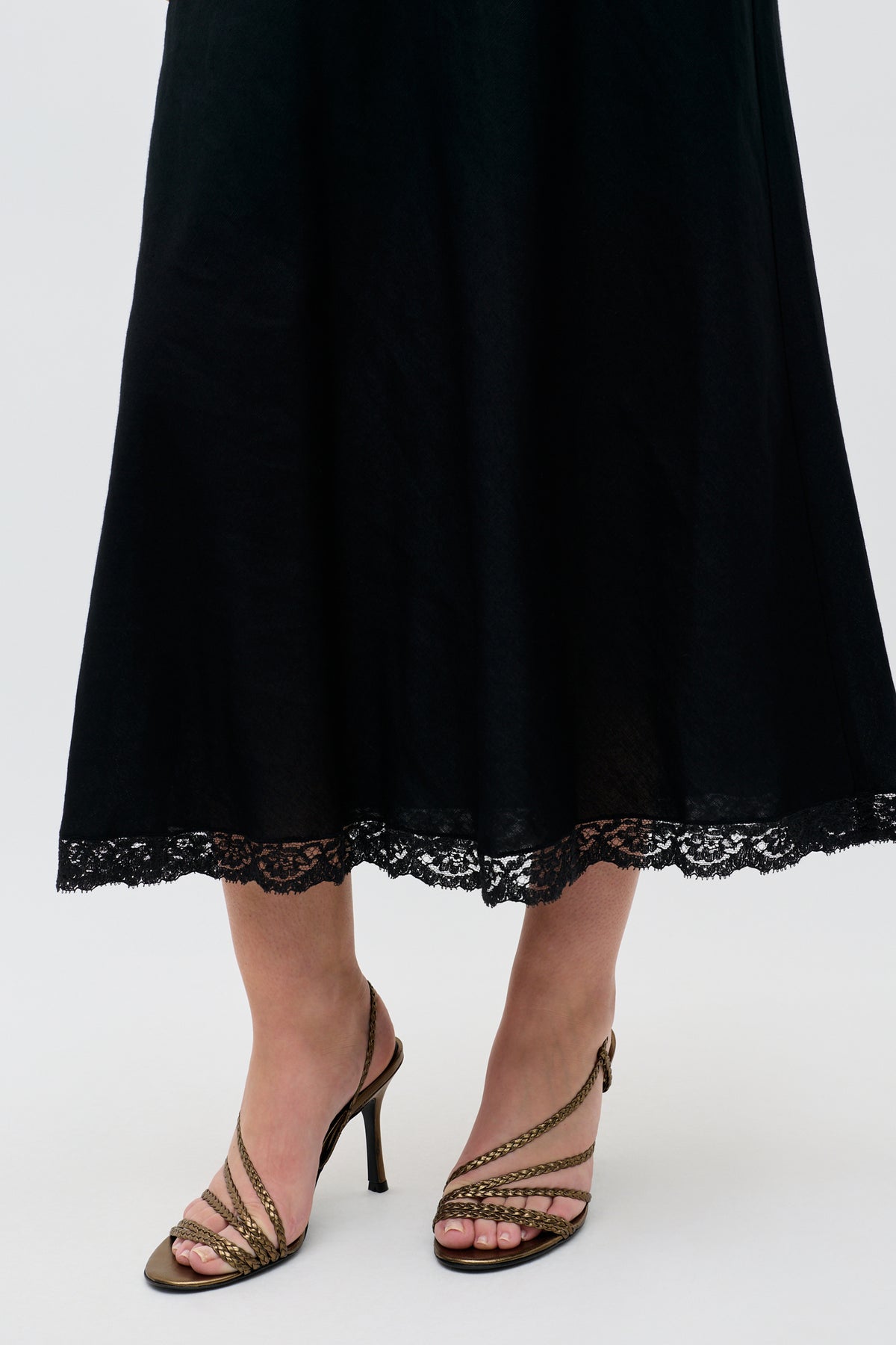 A person wearing a long black skirt with lace trim at the hem and the Gracey - Gold strappy high-heeled sandals standing on a light background.