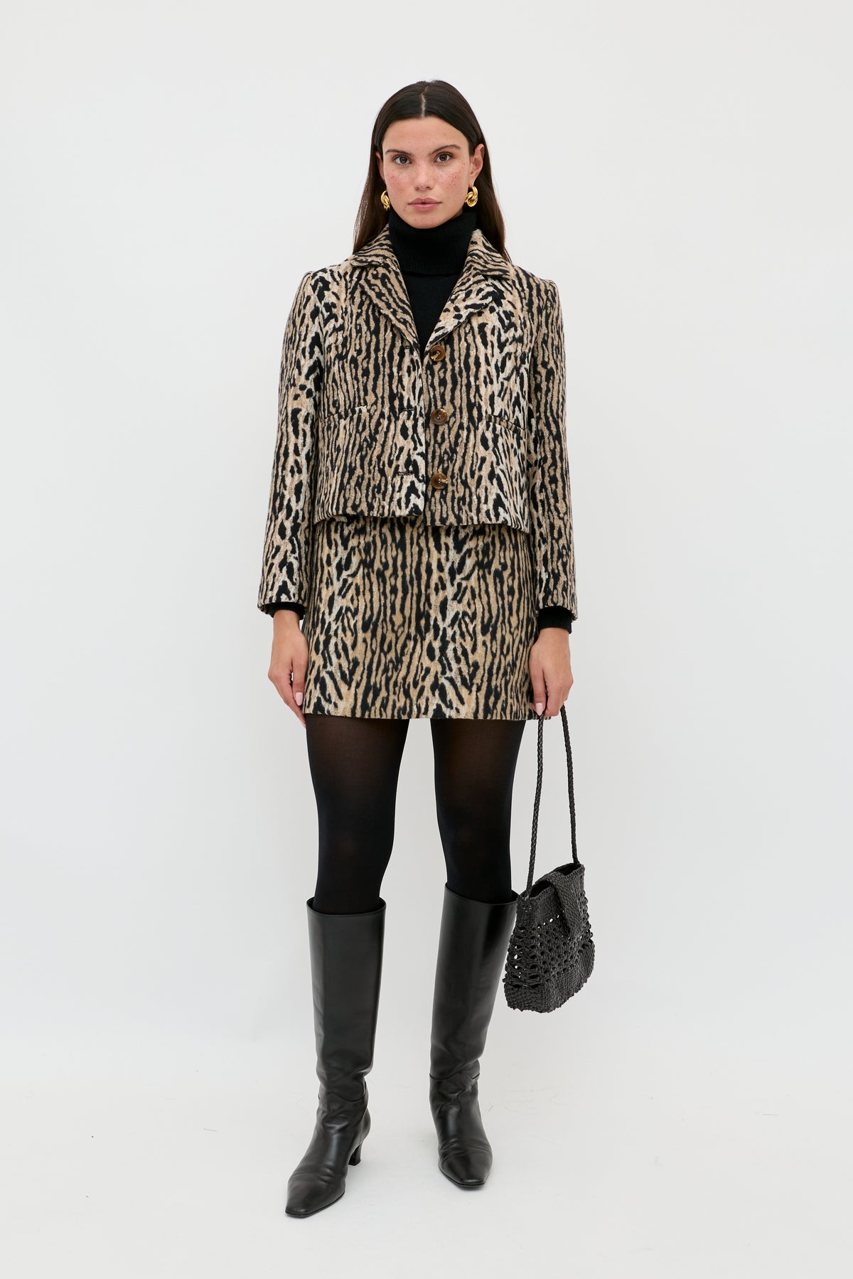 A woman stands against a white background wearing the Leila - Bohemia Leopard Brown jacket and skirt, paired with a black turtleneck, black tights, knee-high black boots, and holding a textured black handbag.