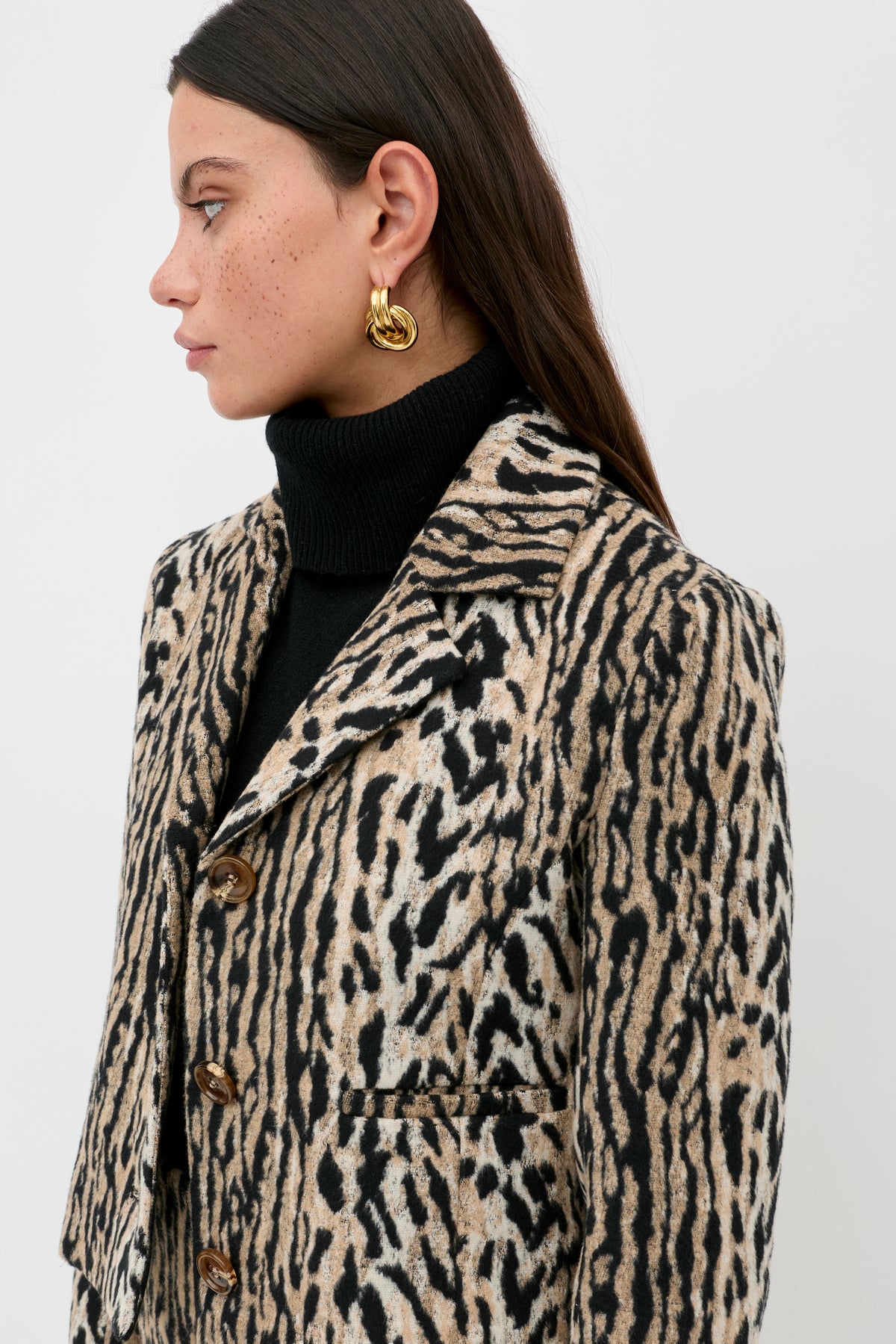 A woman with long dark hair and gold earrings wears an elegant black turtleneck and the Leila - Bohemia Leopard Brown blazer. She is facing left against a plain white background.