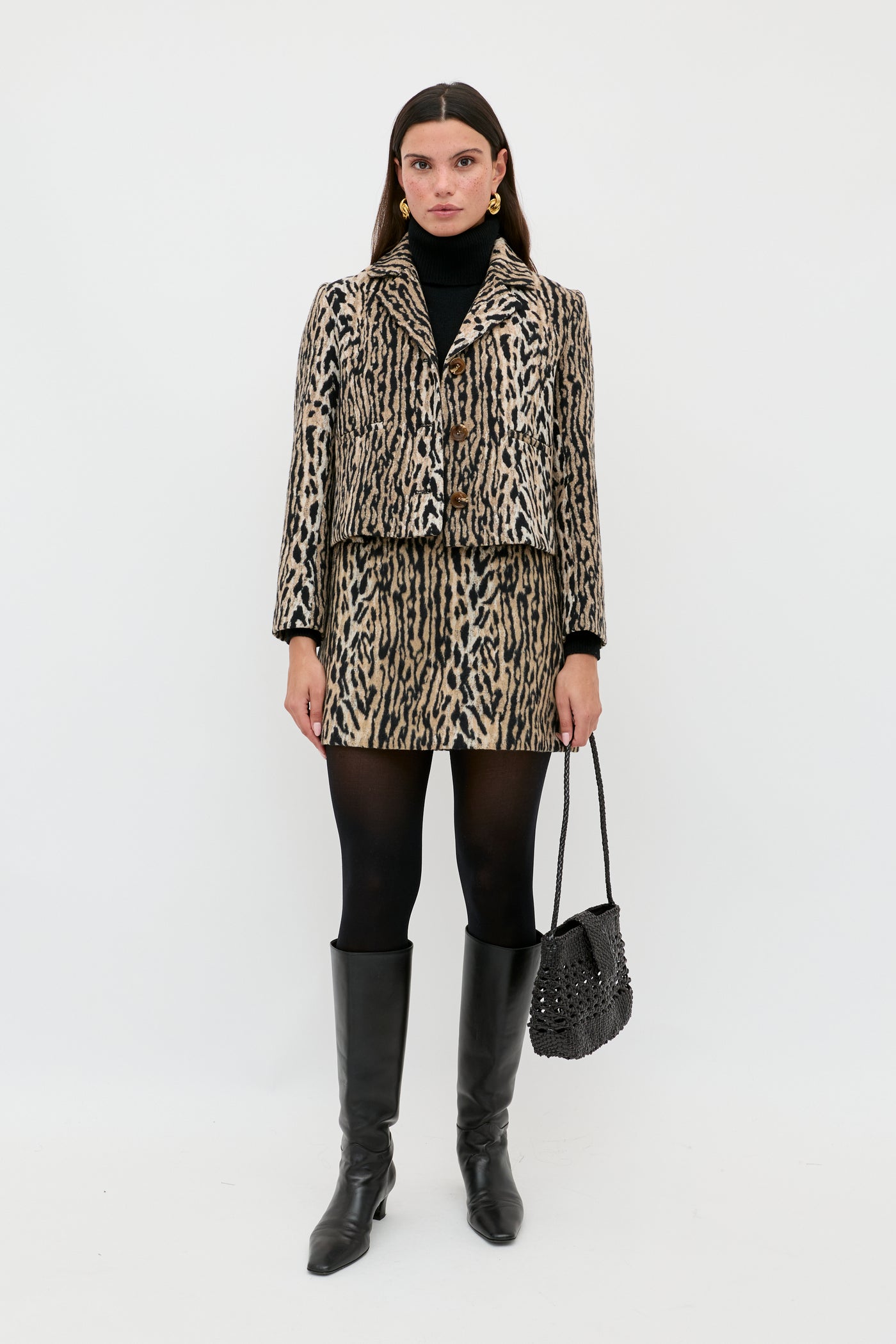 A woman stands against a white background wearing the Leila - Bohemia Leopard Brown jacket and matching skirt with a black turtleneck, tights, knee-high boots, and a black purse, channeling chic vintage flair.