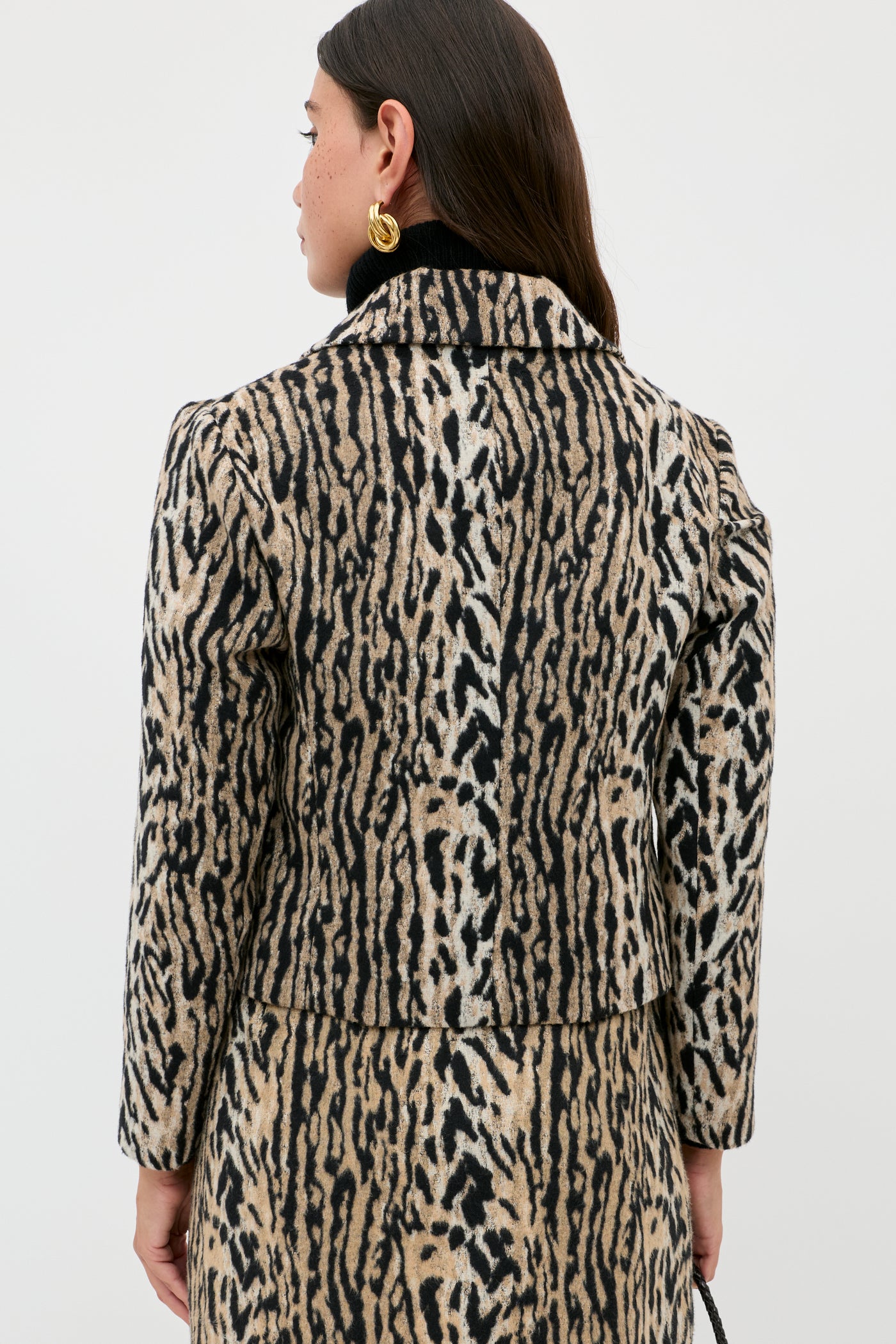 A woman with long dark hair and gold hoop earrings is seen from the back, wearing the Leila - Bohemia Leopard Brown jacket and matching skirt against a plain white background.