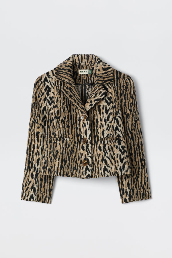 The Leila - Bohemia Leopard Brown is an elegant cropped button-up jacket with a bold beige, brown, and black leopard print, collar, and front pockets. Inspired by vintage souvenir jackets, it's shown on a plain white background.