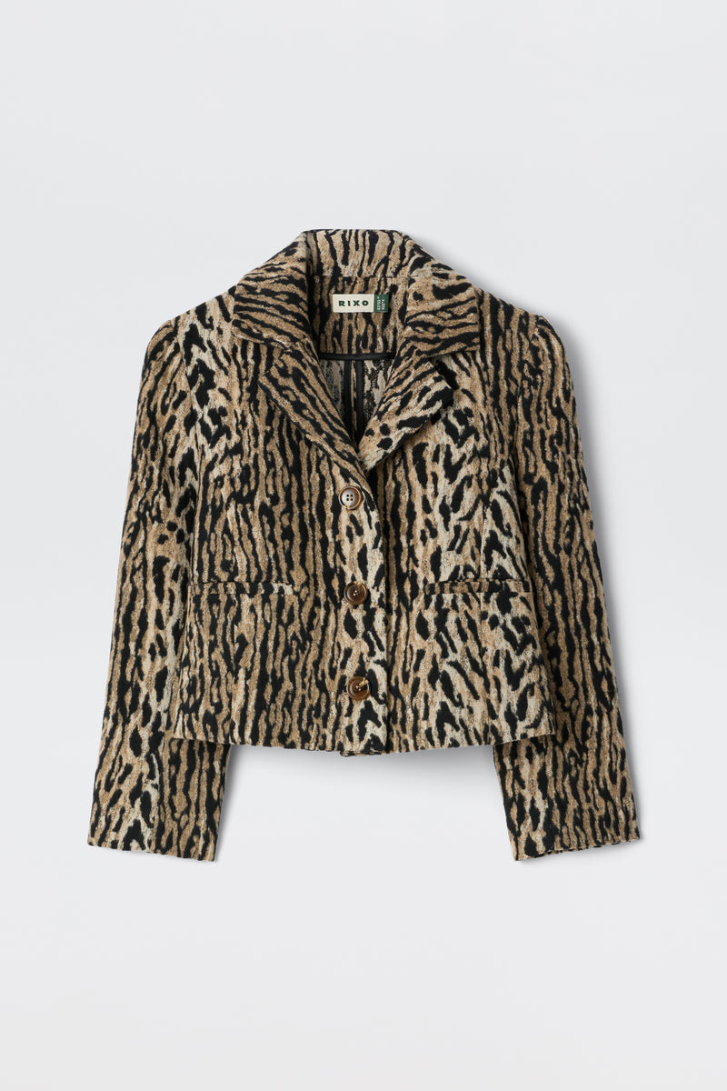 The Leila - Bohemia Leopard Brown is an elegant cropped button-up jacket with a bold beige, brown, and black leopard print, collar, and front pockets. Inspired by vintage souvenir jackets, it's shown on a plain white background.