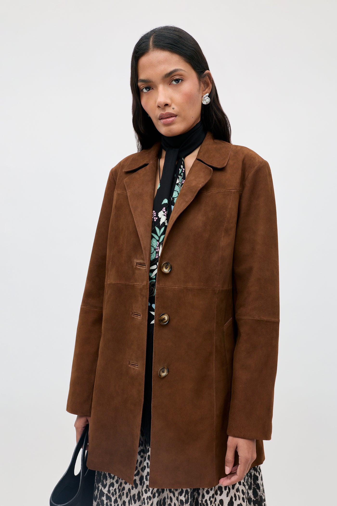 A woman wearing the Lainey - Chestnut suede coat over a floral top and animal print skirt, accessorized with earrings and a black scarf, stands against a plain light background.