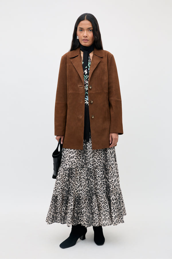 A woman stands facing forward, wearing the Lainey - Chestnut brown suede jacket over a patterned dress with a leopard print skirt, black boots, and holding a small black handbag.